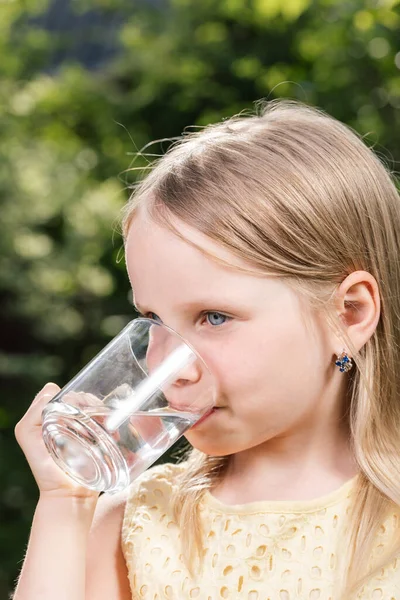 夏の庭のガラスのカップから黄色のドレス飲料水を身に着けている小さな女の子 健康的なライフスタイルの概念 — ストック写真