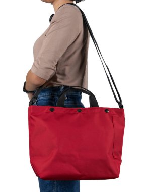 Uzun kollu kahverengi gömlekli ve kotlu bir kadın modelin izole edilmiş yakın çekim stüdyo fotoğrafı beyaz arka planda büyük kırmızı, çok amaçlı uniseks seyahat eden spor çantasından asılı duruyor..