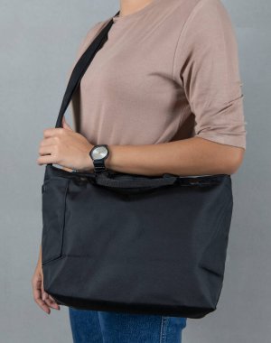 Uzun kollu kahverengi gömlekli ve kotlu kadın modelin izole edilmiş yakın çekim stüdyo fotoğrafı beyaz arka planda büyük siyah, çok amaçlı uniseks seyahat eden spor çantasının asılı duruşu..