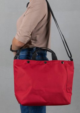 Uzun kollu kahverengi gömlekli ve kotlu bir kadın modelin izole edilmiş yakın çekim stüdyo fotoğrafı beyaz arka planda büyük kırmızı, çok amaçlı uniseks seyahat eden spor çantasından asılı duruyor..