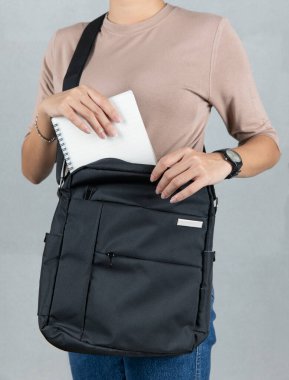 Kahverengi uzun kollu tişörtlü kadın modelin yakın çekim stüdyosu gümüş dizüstü bilgisayarı gri arka planda büyük siyah çapraz gövdeli uniseks çantasının cebine koyuyor..