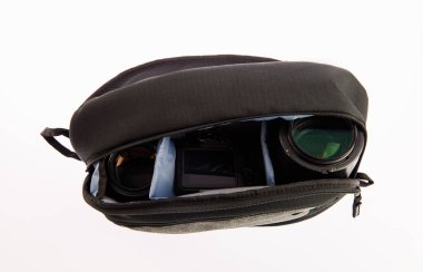 Profesyonel fotoğrafçının, su geçirmez, modaya uygun siyah kumaşlı orta boy sırt çantasının, beyaz arka plandaki yumuşak bölmenin içinde kamera ve lens tutarken çekilmiş yakın çekim görüntüleri..