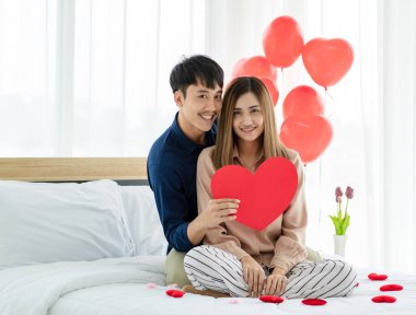 Sevgililer Günü 'nde sevgilisine sarılıp yatakta dinlenirken gülümseyen ve kameraya bakan neşeli Asyalı adam.