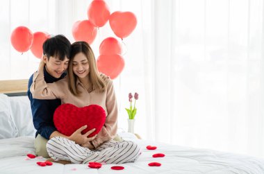 Yastık şeklinde kalp şeklinde gülümseyen ve yatakta birbirini kucaklayan iyimser genç Asyalı çift. Genç aşıkların aşk ve ilişkisi.