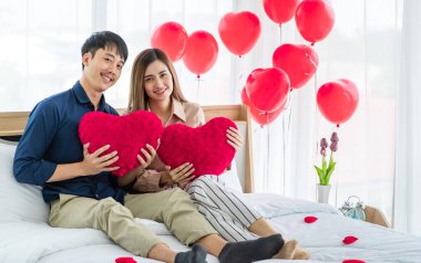 Kalp şeklinde yastıkları olan neşeli Asyalı erkek ve kadın aşıklar Aziz Sevgililer Günü 'nde yatakta dinlenirken gülümseyip kameraya bakıyorlar.