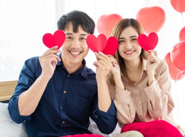 Neşeli Asyalı erkek ve kadın aşıklar Sevgililer Günü kutlamalarında gülümsüyor ve gözlerinin yanında kağıt kalpler tutuyorlar. Mutlu çift aşk konsepti.