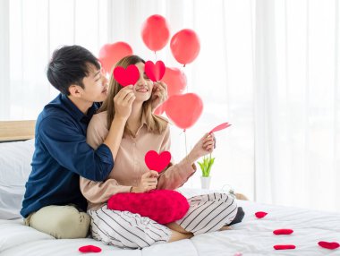 Neşeli Asyalı erkek ve kadın aşıklar Sevgililer Günü kutlamalarında gülümsüyor ve gözlerinin yanında kağıt kalpler tutuyorlar. Mutlu çift aşk konsepti.