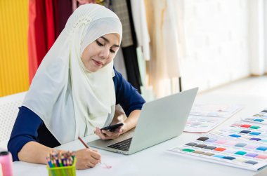 Cep telefonlarını karıştırırken tesettürde gülümseyen ve stüdyoda çalışırken not alan pozitif kadın terzi.