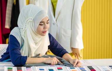 Türbanlı İslami kadın, masa başında oturmuş terzilik atölyesinde çalışırken kumaş örnekleriyle karşılaştırıyor.