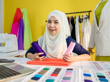 Türbanlı İslami kadın, masa başında oturmuş terzilik atölyesinde çalışırken kumaş örnekleriyle karşılaştırıyor.