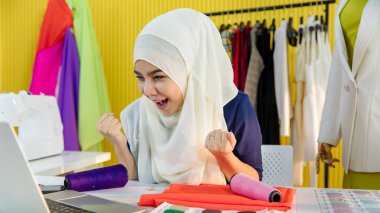 Mutlu İslami kadın terzi yumruğunu sıkıca kavrayıp bağırırken dizüstü bilgisayar kullanıyor ve atölyedeki başarıyı kutluyor.