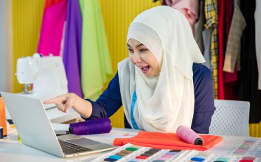 Mutlu İslami kadın terziler bilgisayar ekranını işaret ederek dizüstü bilgisayar kullanırken ve atölyede başarıyı kutlarken heyecanlanır.