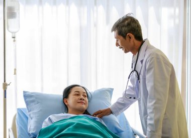 Stetoskop monitörüyle beyaz önlüklü yaşlı Asya 'lı gri saçlı ve bıyıklı doktor. Normal salin çözeltisi damlalarıyla hastane yatağında yatan kadın hastaların kalp atışlarını dinle..