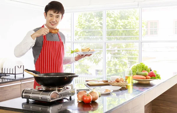 Asyalı yakışıklı kocanın portre resmi kırmızı çizgili önlük giyiyor gülümsüyor mutfak tezgahında yemek pişirirken kameraya bakıyor.