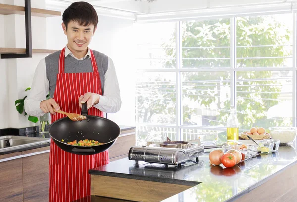 Asyalı yakışıklı kocanın portre resmi kırmızı çizgili önlük giyiyor gülümsüyor mutfak tezgahında yemek pişirirken kameraya bakıyor.