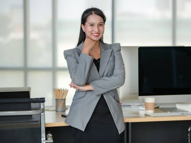 Gri takım elbiseli genç çekici Asyalı kadın bulanık pencereli modern ofis özgüveniyle ayakta duruyor. Modern ofis yaşam tarzı kavramı.