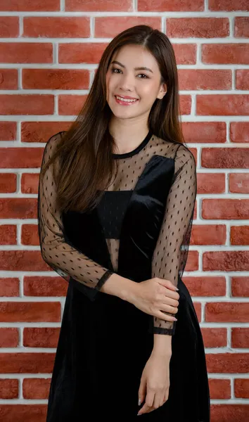 年轻美丽迷人的亚洲女模特形象 身披黑衣 面带微笑地站在橙色砖墙的背景下 — 图库照片