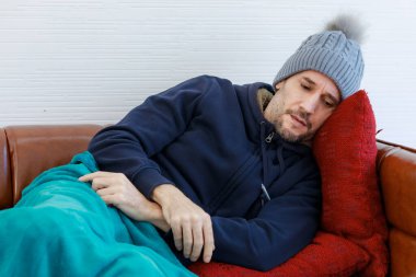 Kafkasyalı hasta, sakallı, yün ceketli, deri koltuğa ve yastığa uzanmış, battaniyeyle sarılmış, Coronavirus salgınında baş ağrısı ve soğuk algınlığı olan bir adam..