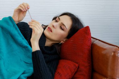 Yün kazaklı hasta beyaz bir kadının yakın plan fotoğrafı yastık ve deri koltuğa uzanmış battaniye tutarak termometreyi elinde tutarak kışın grip belirtileri gösteren yüksek ısıyı ölçüyor..