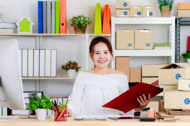Asyalı genç, mutlu, çevrimiçi iş kadını satıyor. Müşteri adreslerini bilgisayar monitörü ve klasöründen kontrol ediyor. Çalışma masası karton karton kargo kutularıyla dolu..