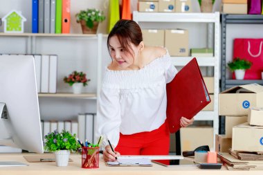 Asyalı başarılı çevrimiçi satıcı iş kadını elinde klasör tutarken ofis ofisindeki karton kutu tablet bilgisayar monitörüyle dolu çalışma masasında kağıt tahtadaki müşteriden kalem siparişi kullanıyor..