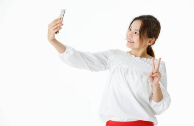 Asyalı mutlu kadın modelin gündelik uzun kollu gömleğiyle tek başına çekilmiş bir fotoğrafı. İki parmak zafer işareti var. Akıllı telefon kamerasına bakıyor ve beyaz arka planda selfie çekiyor..