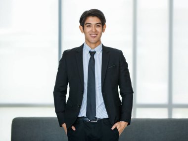 Resmi bir takım elbise giyen yakışıklı ve başarılı Asyalı iş adamı portresi. Sırıtarak gülümsüyor, ellerini ceplerine sokuyor, güvenli ve akıllı bir şekilde ayakta duruyor ve kapalı ofis kapısı açık.