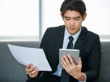 20 'li yaşlarda yakışıklı, genç Asyalı bir işadamı kravat takıyor, oturuyor ve finansal pazarlama raporlarını okuyor.