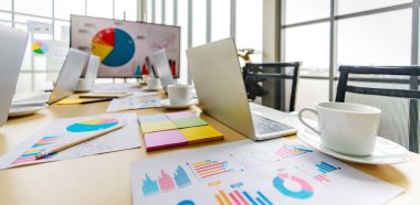 Çalışma masasına dizüstü bilgisayarla yerleştirilen küçük tabağa konmuş beyaz, lezzetli kahve fincanı ön hedef büyüme hızı grafiği ve arka plandaki büyük ekran monitörünün çizelgesi..