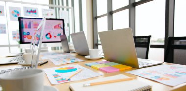 Bilgisayarın renkli dizüstü bilgisayarı, kağıt rapor grafiği dokümanları konferans salonundaki masanın üzerine yerleştirilmiş analiz satış hedefleri ekranda ve cam panoda bulanık arka planda..