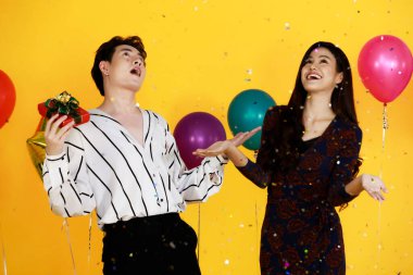 Asyalı mutlu erkek ve kadın aşığın stüdyo fotoğrafları şık giyinen çift, sarı arka planda balonlar olan kutlama partisinde parlak kağıt konfetiyle eğleniyor..
