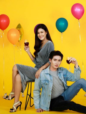 Asyalı mutlu erkek ve kadın aşığın stüdyo fotoğrafları şık giyinmiş çift kırmızı şarap kadehleriyle oturmuş kutlama partisinde kameraya poz veriyor sarı arka planda balonlar var..