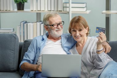 Beyaz, yaşlı, yaşlı, yaşlı bir çift. Oturma odasındaki kanepede birbirlerine sarılıyorlar. Kredi kartı ve dizüstü bilgisayarla online mağazadan internet alışverişi yapıyorlar..