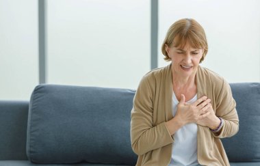 Beyaz, yaşlı, yaşlı, yaşlı, sağlıksız hasta kadın ev hanımı. Kanepede tek başına oturuyor. Göğsünde el ele tutuşup acil kalp krizi geçiriyor..