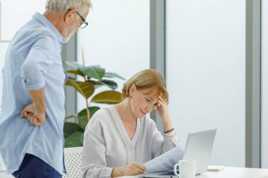 Beyaz, yaşlı, yaşlı, yaşlı, bunalımlı kadın ev hanımının mali tasarruf verileri okuması endişesi baş ağrısı. Gri sakallı koca ise bulanık ön planda destek veriyor..