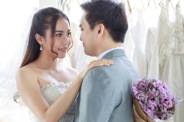 Gri takım elbiseli Asyalı yakışıklı damat beyaz uzun gelinlik içinde güzel mutlu bir gelinle soyunma odasında el ele tutuşup çiçek buketini birlikte taşıyor..