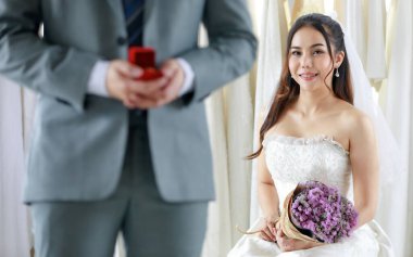 Gri takım elbiseli tanınmayan damat kırmızı elmas yüzük kutusunu tut ve Asyalı genç ve mutlu geline beyaz gelinlik içinde bulanık arka planda çiçek buketi ver..