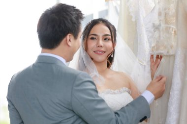 Beyaz tüylü dantel peçeli, Asyalı güzel gelin. Elinde gülümsemeyle diğer kıyafetlere bakıyor. Soyunma odasında gri takım elbiseli yakışıklı damatla birlikte..