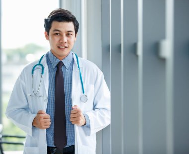 Üniformalı, neşeli, genç ve yakışıklı Asyalı erkek doktor ve steteskop klinikte dikilip dışarıya bakıyor.