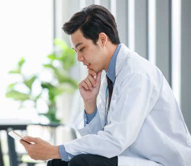 Hastanede tıp odasında oturan ve cep telefonlarını karıştıran meşgul Asyalı erkek pratisyen doktorun yan görüntüsü