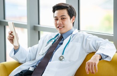 Hastanede tıp odasında oturan ve cep telefonlarını karıştıran meşgul Asyalı erkek pratisyen doktorun yan görüntüsü