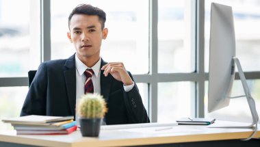 Mutlu yüzü ve kendine güveni olan, resmi takım elbiseli Asyalı genç erkek girişimci masaüstü bilgisayarının yanında oturuyor..