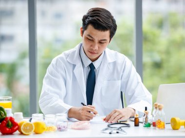 Tıp üniformalı Asyalı bir adam modern klinikte çalışırken hap alıyor ve not alıyor.