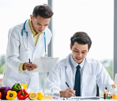Tıp üniformalı Asyalı erkek meslektaşlar modern klinikte çalışırken meyvelerle birlikte masa üzerinde birlikte çalışıyorlar.
