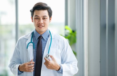Hastanede çalışırken kameraya gülümseyen, tıbbi üniformalı mutlu Asyalı genç doktor portresi.