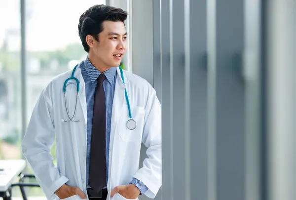 제복을 청진기를 쾌활하고 귀여운 아시아 의사가 진료소의 주머니에 바깥을 바라보고 — 스톡 사진