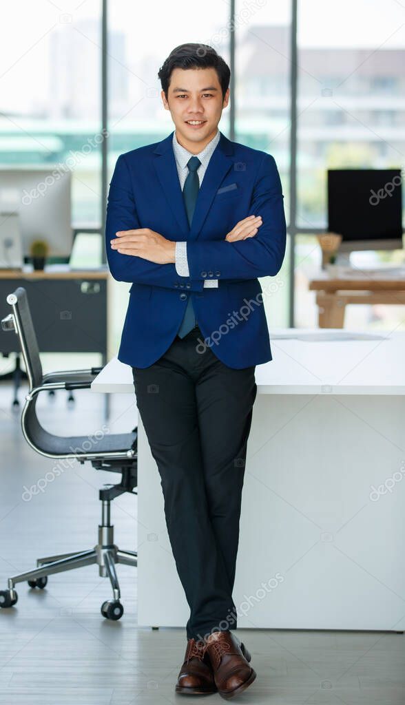 Confident Asian male entrepreneur in formal suit standing with crossed arms in modern workplace and looking at camera