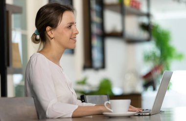 Beyaz uzun kollu gömlekli güzel beyaz kadın öğleden sonra kafede rahat bir şekilde oturuyor ve bilgisayarında online iş için daktilo yazıyor..