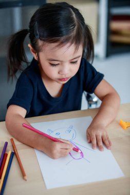 Küçük güzel at kuyruğu saç stili anaokulu kızı sandalyeye oturmuş, resim çiziyor, renkli kalemleri kağıt üzerine çiziyor evdeki oturma odasındaki kitaplığın önündeki masaya..
