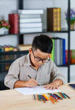 Gözlüklü ve gri tişörtlü zeki Asyalı çocuk okuma odasında tablet, cep telefonu ve renkli kalemlerden oluşan çalışma masasında oturuyor ve beyaz kağıda konsantre oluyor.
